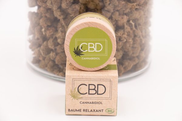 Packaging Baume Relaxant CBD Bio MONPLANCBD devant un grand bocal de fleurs de CBD (décor).
