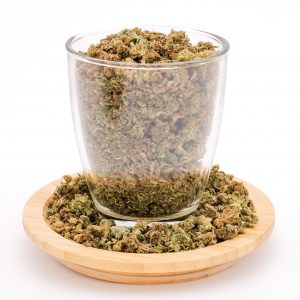 Clear glass filled with MIX SMALL BUD + TRIM cannabis, sitting on wooden plate with more blend scattered around.