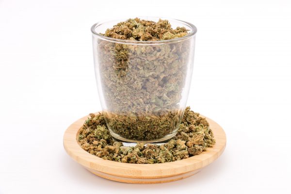 Clear glass filled with MIX SMALL BUD + TRIM cannabis, sitting on wooden plate with more blend scattered around.