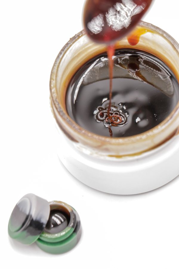 Rosin CBD liquide premium MONPLANCBD (61.83% CBD) coulant d'un outil dans un pot en verre, avec un petit contenant silicone à côté - Achat CBD en ligne.