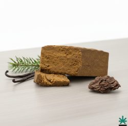Gros plan sur la texture sableuse du A6 Dry Sift Hash CBD, avec une gousse de vanille et une pomme de pin en arrière-plan.